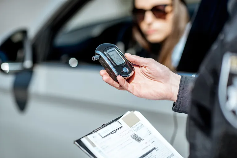 Police officer holding a breathalyzer device near a woman sitting inside a white car.