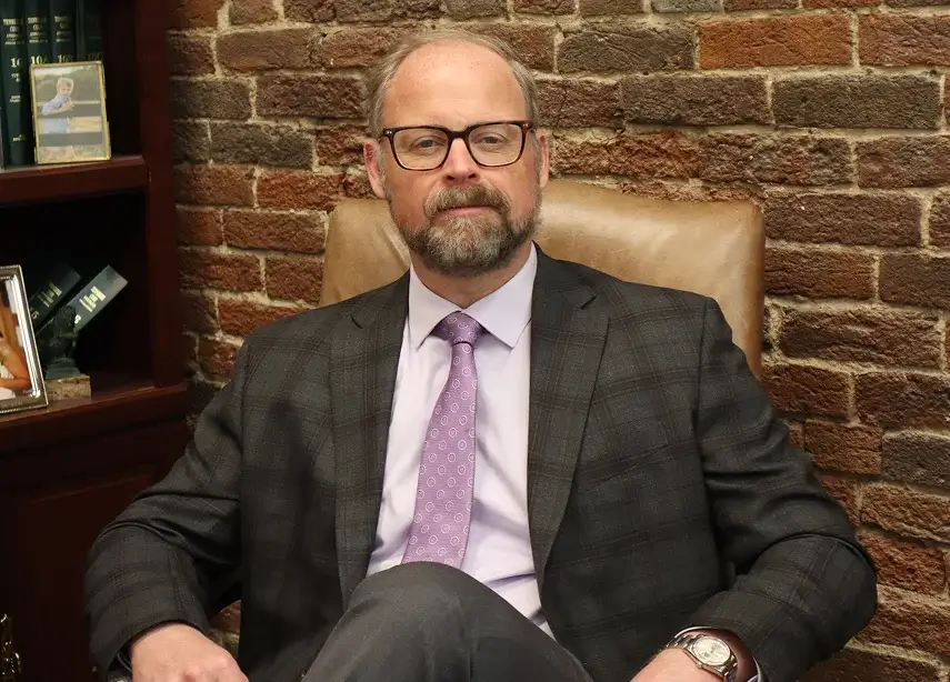 Erik Herbert wearing glasses and a suit sits confidently in an office chair, surrounded by a professional office environment