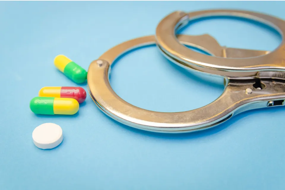 Metallic handcuffs placed beside multicolored capsules and a white round tablet on a blue surface.
