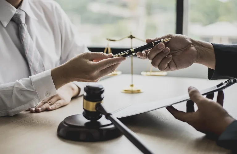 Two professionals exchanging a pen across a desk with a gavel and legal scales in the background.