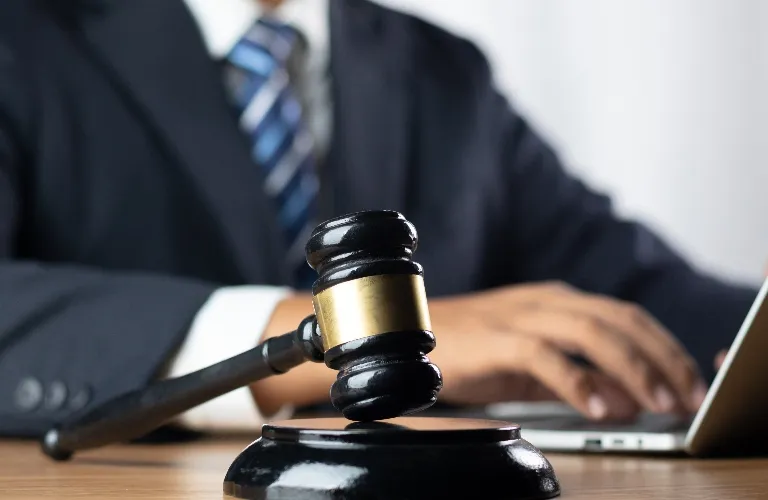 Close-up of a black judge's gavel with brass band on wooden desk, blurred person in suit typing on laptop.