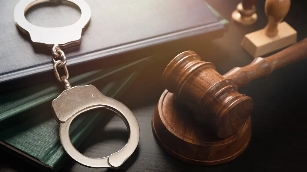 Wooden judge's gavel, metal handcuffs, and stacked law books on a dark surface.