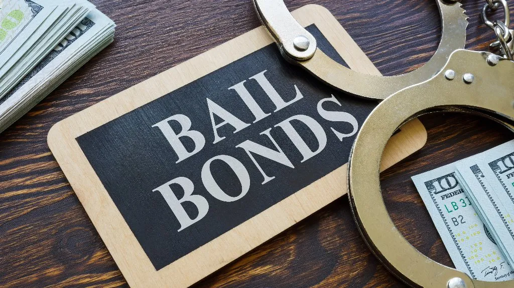 Wooden sign reading "BAIL BONDS" next to metal handcuffs and stacks of US hundred-dollar bills on a wooden surface.