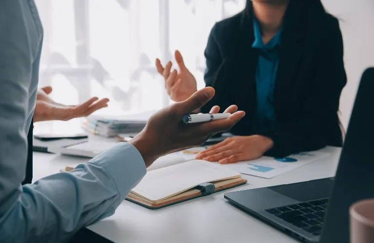 Two professionals engaged in discussion at a desk with notebook, laptop, and documents.