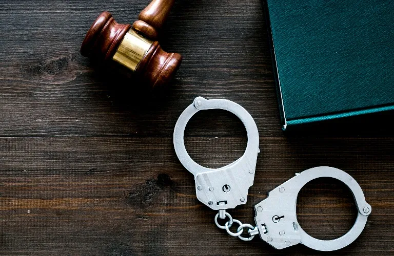 Wooden judge's gavel, metal handcuffs, and closed green book on dark wooden surface.
