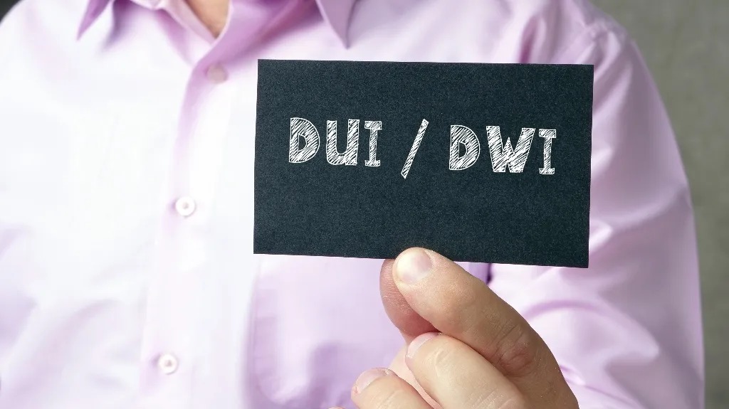 Person in light purple shirt holding black card with white text "DUI / DWI" in focus.