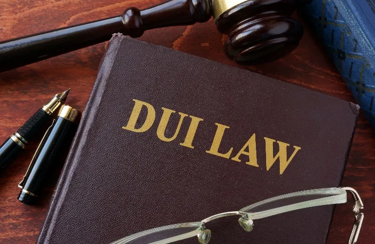 Close-up of a brown book titled "DUI LAW" with a gavel, fountain pen, and eyeglasses on a wooden surface.