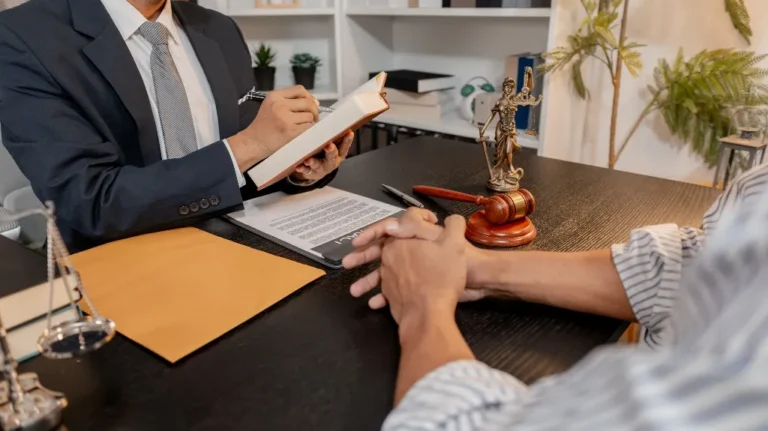 Two individuals in a legal consultation, with a gavel, Lady Justice statue, and documents on a dark desk.