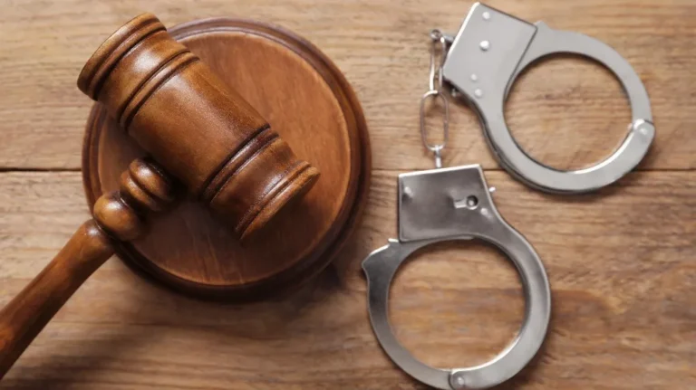 Wooden judge's gavel and round sound block beside metal handcuffs on wooden surface.