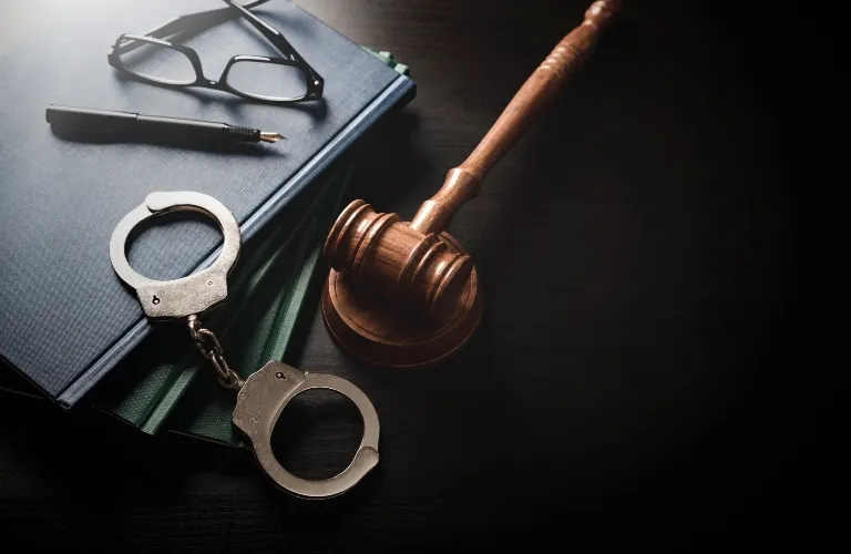 Wooden judge's gavel, metal handcuffs, eyeglasses, and stacked books on dark surface.