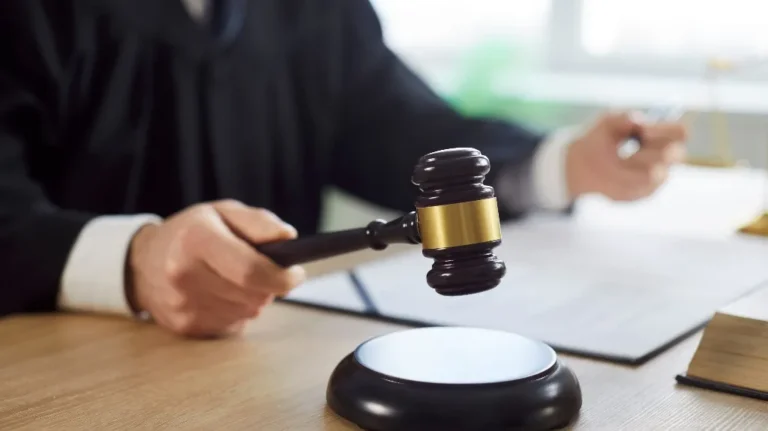 Judge in black robe holding wooden gavel above sound block on desk.