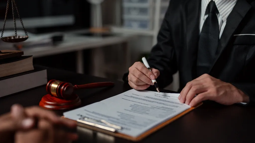 Person in black suit signing legal documents at desk with gavel and scales of justice.