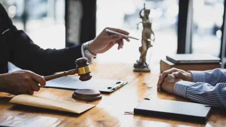Two individuals at a wooden table with a judge's gavel, legal documents, and a Lady Justice statue.