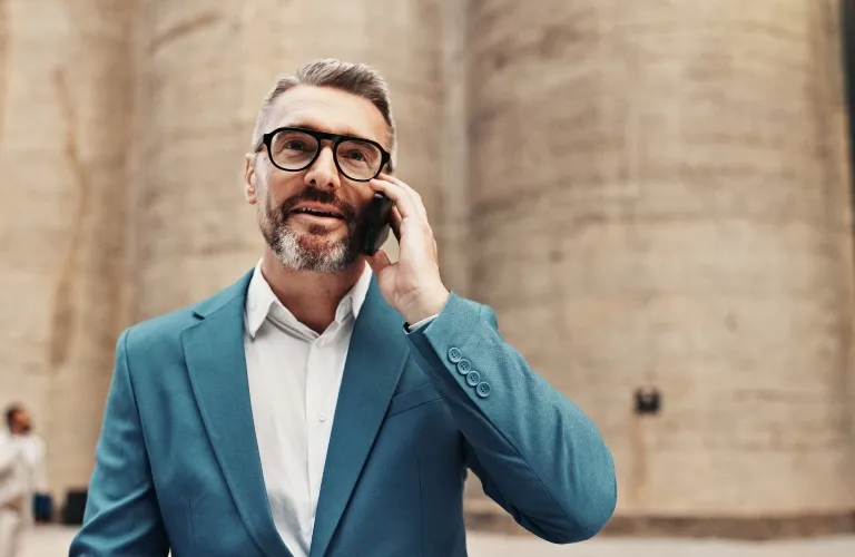 Middle-aged man in teal blazer and glasses speaking on smartphone, neutral background.