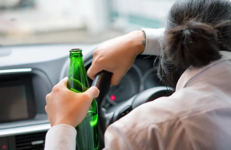 Person holding a green glass bottle while gripping a car steering wheel, viewed from behind.