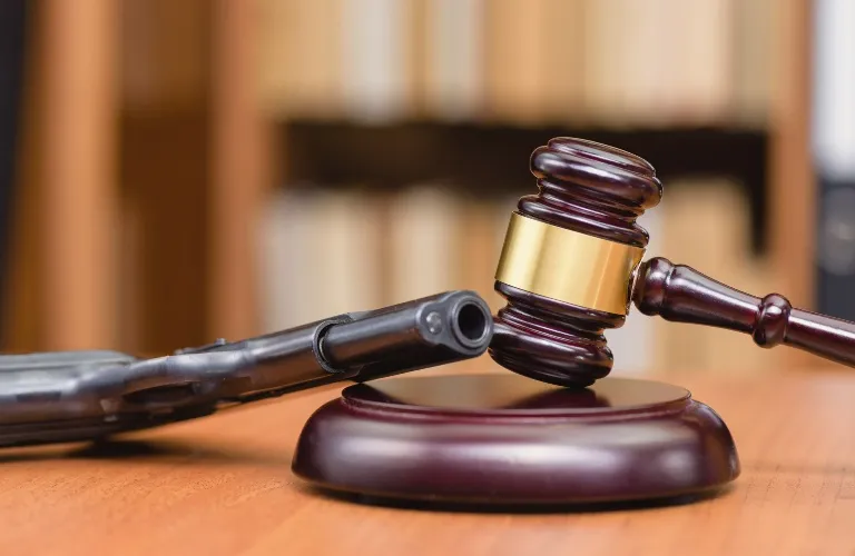 Wooden judge's gavel with gold band next to black handgun on wooden table.