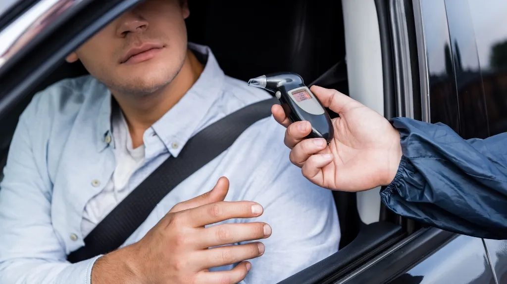 A man in a light blue shirt sitting in a car is offered a breathalyzer device by an officer.
