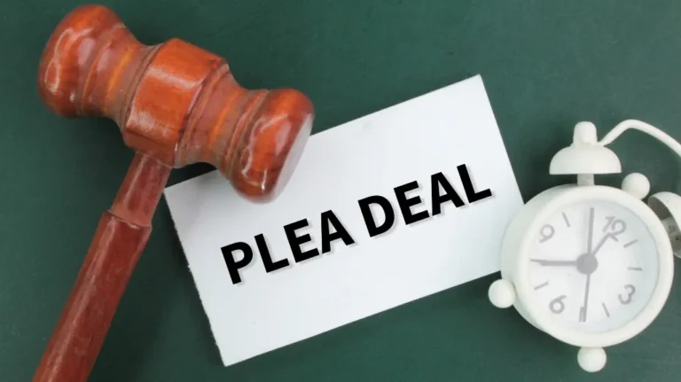 Wooden judge's gavel, white card with "PLEA DEAL" text, and white alarm clock on green surface.