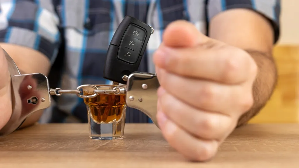 Person wearing handcuffs with a car key and a shot glass of alcohol on a wooden table.