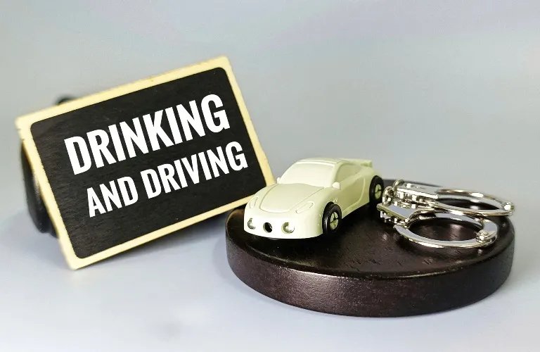 A small white toy car and metal handcuffs on a wooden base, with a sign reading "DRINKING AND DRIVING.