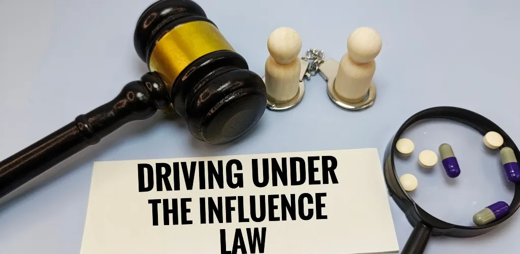 Wooden judge’s gavel, two wooden figurines with handcuffs, magnifying glass with pills, and a paper stating "Driving Under The Influence Law".