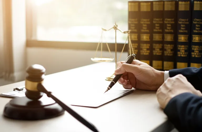Hand holding pen over document on desk with gavel, scales of justice, and legal books.