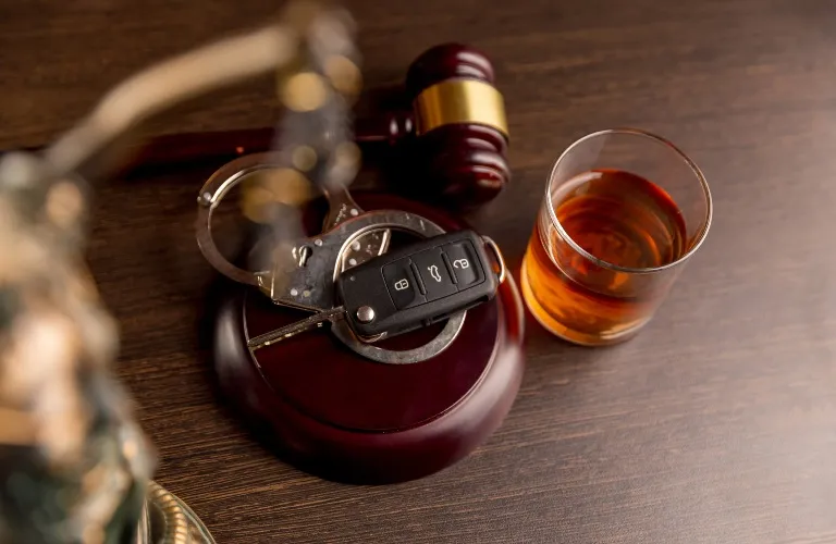 Wooden judge's gavel with handcuffs, car key, and a glass of amber liquid on a dark surface.