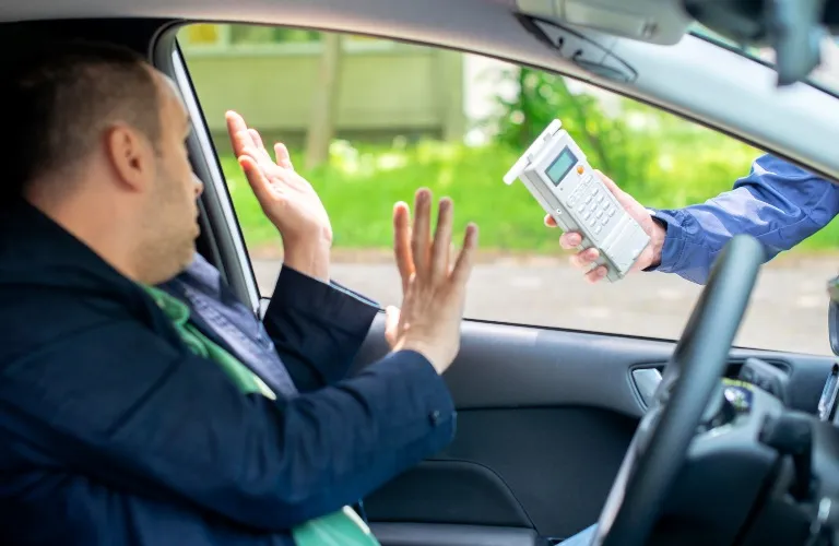 Man inside car raising hands as police officer extends breathalyzer device.