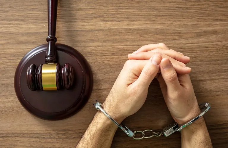 Hands in silver handcuffs resting on a wooden table next to a dark wooden judge's gavel.