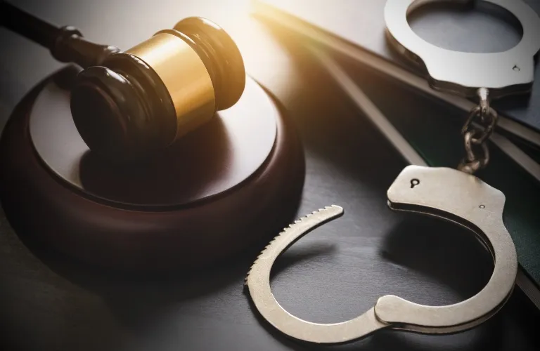 Wooden judge's gavel and metallic handcuffs on a dark surface with sunlight.