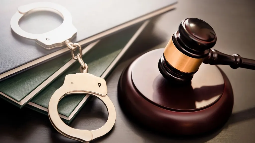 Wooden judge's gavel with brass band next to silver handcuffs and stacked law books.