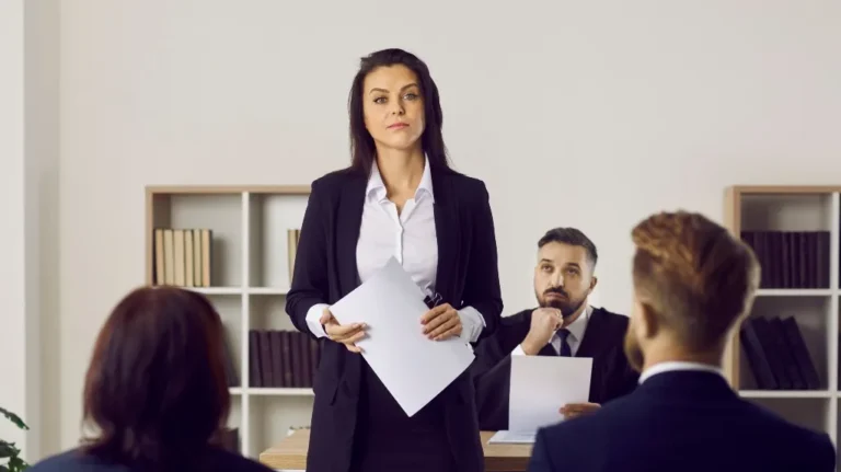 Professional woman in black suit presenting documents to seated colleagues in office setting.