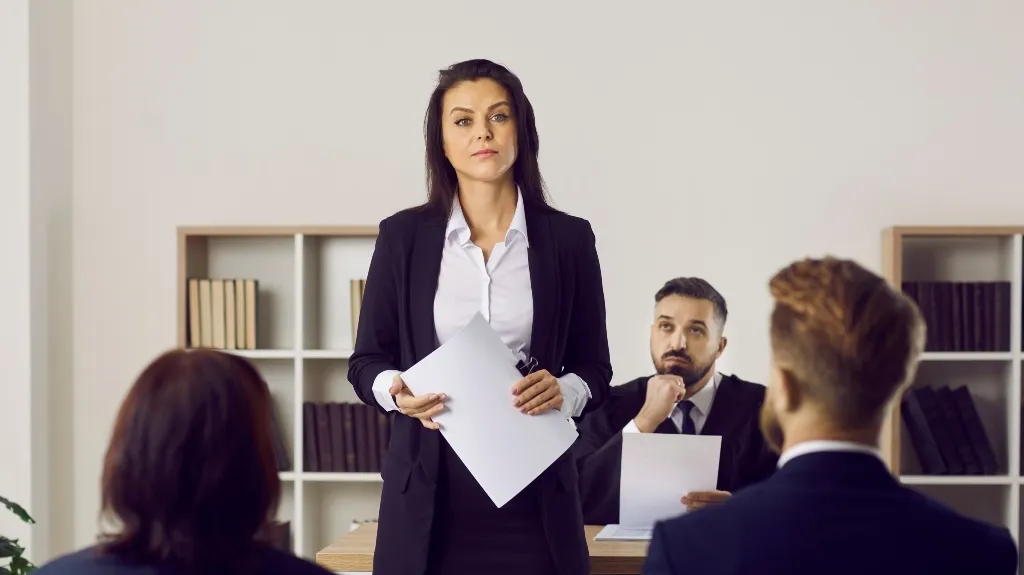 Professional woman in black suit presenting documents to seated colleagues in office setting.