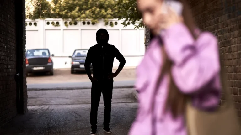 Silhouetted person in hoodie standing in alleyway, blurred woman in purple jacket on phone in foreground.