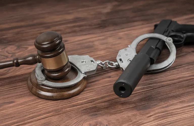 Wooden judge's gavel, silver handcuffs, and black silenced handgun on wooden surface.