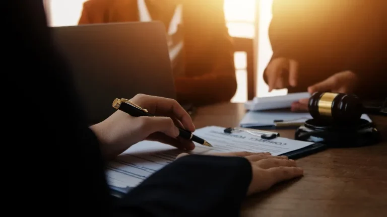 Person signing a contract document at a wooden table with a gavel and laptop nearby.